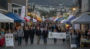 A vibrant local event on Bainbridge Island with small business booths, community members walking, banners, and signage promoting local vendors. Include warm evening lighting, people taking photos